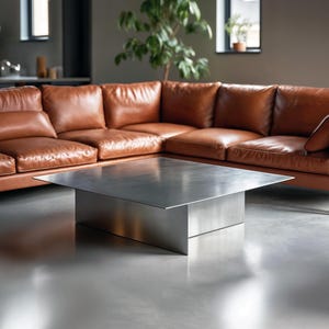 Peut inclure: Une table basse contemporaine avec une finition en métal brossé. La table a un plateau rectangulaire et une base solide en forme de boîte. Un grand canapé d'angle en cuir marron est en arrière-plan.