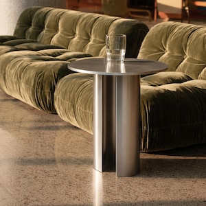 Może przedstawiać: Nowoczesny stolik boczny ze szczotkowanego metalu z okrągłym blatem i cylindryczną podstawą. Na stole stoi szklanka czystej wody. Za nim duża, oliwkowozielona aksamitna sofa. Podłoga jest nakrapiana.