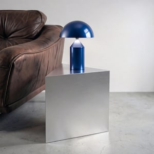 Può includere: Una moderna lampada da tavolo blu navy, con un paralume arrotondato e una base cilindrica, è posizionata su un tavolino laterale in metallo spazzolato. La lampada è accesa, emettendo una luce soffusa. Una poltrona in pelle marrone è parzialmente visibile sullo sfondo.