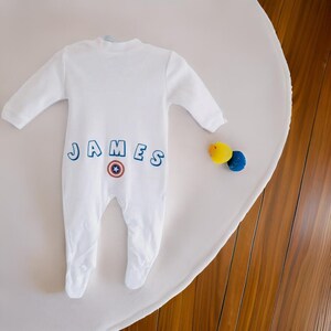 Könnte beinhalten: Weißer Baby-Strampler mit dem Namen "JAMES" in blauen umrandeten Buchstaben und einem rot-weiß-blauen Captain America-Logo auf der Rückseite.
