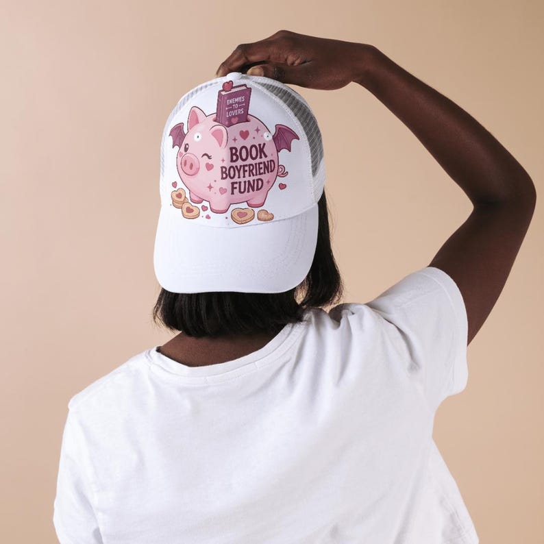 May include: A white trucker hat featuring a graphic of a pink winged piggy bank with the text "BOOK BOYFRIEND FUND" and a small book labeled "ENEMIES TO LOVERS". The cap has a solid white front panel and a white mesh back panel with a curved brim.