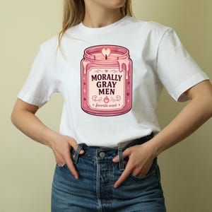 May include: A white short-sleeved t-shirt featuring a graphic of a pink candle in a glass jar. The label on the jar reads MORALLY GRAY MEN and favorite scent in dark text, surrounded by small heart and star icons with a heart-shaped flame.