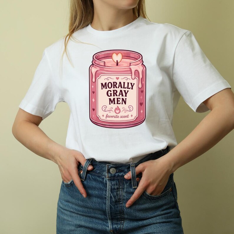 May include: A white short-sleeved t-shirt featuring a graphic of a pink candle in a glass jar. The label on the jar reads MORALLY GRAY MEN and favorite scent in dark text, surrounded by small heart and star icons with a heart-shaped flame.