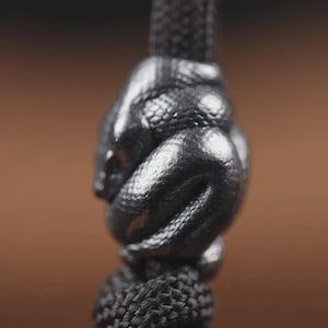 May include: A close-up of a black metal snake pendant coiled around a black cord. The snake has detailed scales and a textured surface. The background is a blurred brown.