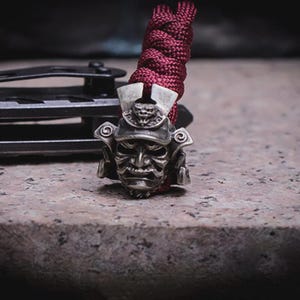 May include: A detailed silver-coloured samurai helmet bead, attached to a burgundy braided cord. The helmet features an intricate face and ornate headgear. The bead is likely used for a keychain or paracord project.