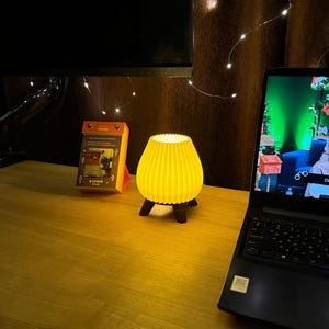 May include: A small, yellow, ribbed lamp with a rounded shape and three dark legs sits on a wooden desk. A laptop and a calendar are also on the desk. The lamp is illuminated, casting a warm glow.