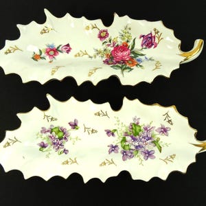 Peut inclure: Deux plats de service en porcelaine en forme de feuille. Les plats sont blancs avec des bords festonnés et une bordure dorée. Chaque plat est décoré d'une composition florale dans les tons de rose, de violet et de jaune.