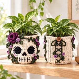 Könnte beinhalten: Zwei cremefarbene, gehäkelte Übertöpfe mit grünen Pflanzen. Ein Übertopf zeigt ein schwarzes Totenkopfgesicht mit violetten Blumen. Der andere hat einen braunen Knopf, eine grüne Schnürung und kleine violette Blüten.