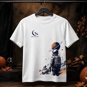 Pode incluir: Uma t-shirt branca de manga curta com um gráfico de um astronauta em pé numa superfície rochosa a olhar para um planeta com anéis. A parte superior esquerda do peito apresenta um ícone de lua crescente com um satélite e o texto ORBIT DREAMER.