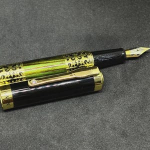 May include: A black and gold fountain pen with a decorative design on the barrel. The pen has a gold nib and a gold clip. The cap is partially removed, revealing the pen's design.