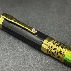 May include: Close-up of a black and gold fountain pen with a green and gold patterned barrel. The pen has a gold clip and a small jewel. The word "PIKA" is visible on the pen.