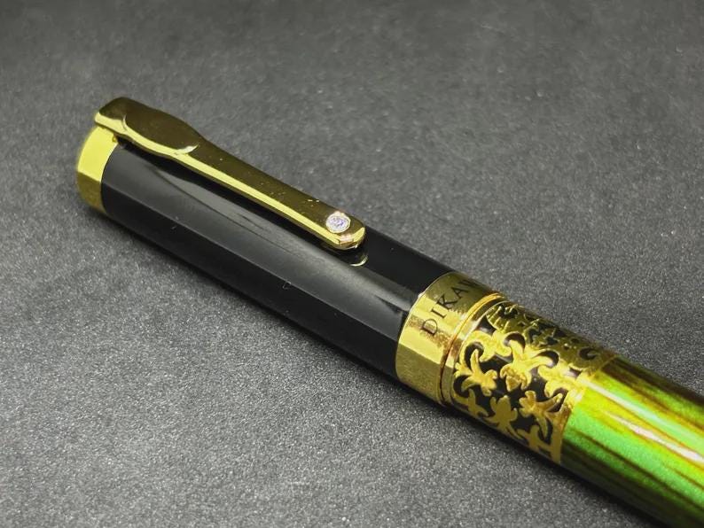 May include: Close-up of a black and gold fountain pen with a green and gold patterned barrel. The pen has a gold clip and a small jewel. The word "PIKA" is visible on the pen.