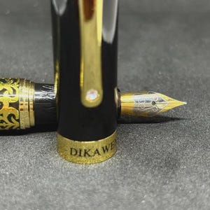 May include: A black and gold fountain pen with a gold nib. The pen has a black barrel with gold accents and a gold cap. The nib is engraved with a decorative design. The pen's cap is off, revealing the nib.
