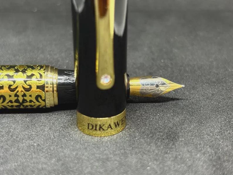 May include: A black and gold fountain pen with a gold nib. The pen has a black barrel with gold accents and a gold cap. The nib is engraved with a decorative design. The pen's cap is off, revealing the nib.