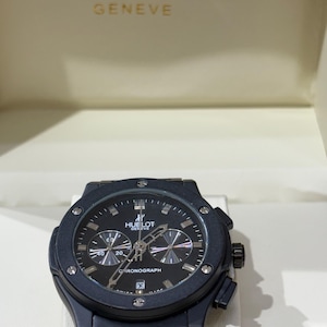 May include: A black Hublot Geneve chronograph watch with a round bezel featuring six visible screws, a black dial with silver-toned hands, three sub-dials, a date window at the six o'clock position, and a black strap, presented in a branded box.