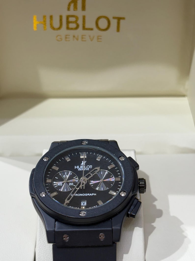 May include: A black Hublot Geneve chronograph watch with a round bezel featuring six visible screws, a black dial with silver-toned hands, three sub-dials, a date window at the six o'clock position, and a black strap, presented in a branded box.