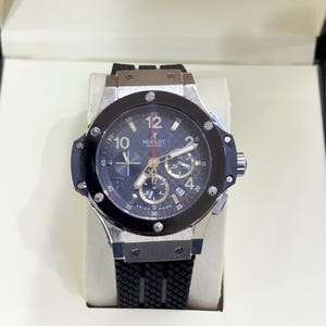 May include: A Hublot Geneve chronograph wristwatch with a black textured rubber strap, a silver-tone metal case, and a black bezel secured by six screws. The black dial features a checkered pattern, luminous hands, sub-dials, and the text Hublot Geneve.