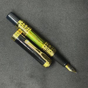 May include: A black and gold fountain pen with a decorative design on the barrel and cap. The pen has a gold nib and is lying on a dark surface. The cap is off, revealing the pen's ink chamber.