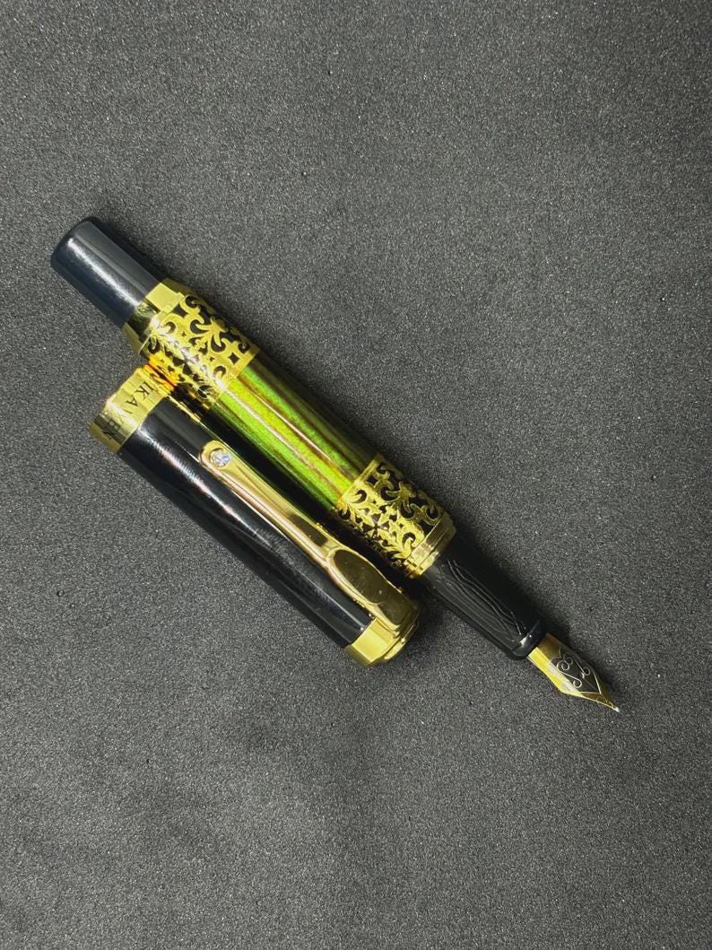 May include: A black and gold fountain pen with a decorative design on the barrel and cap. The pen has a gold nib and is lying on a dark surface. The cap is off, revealing the pen's ink chamber.