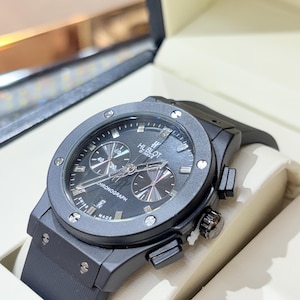 May include: A black Hublot Geneve chronograph watch with a round matte black bezel secured by six silver-toned screws. The black dial features silver hour markers, three sub-dials, and the text Hublot Geneve and Chronograph. It has a black rubber strap.