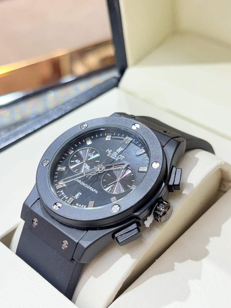 May include: A black Hublot Geneve chronograph watch with a round matte black bezel secured by six silver-toned screws. The black dial features silver hour markers, three sub-dials, and the text Hublot Geneve and Chronograph. It has a black rubber strap.