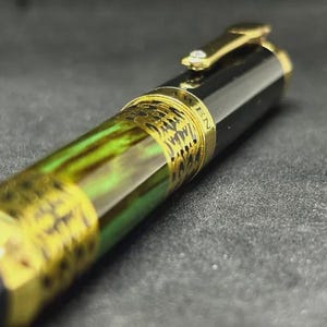 May include: Close-up of a luxury fountain pen with a black barrel and gold accents. The pen features a decorative band with intricate detailing and a green and gold marbled section. The clip is gold with a small gem.