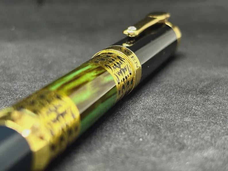 May include: Close-up of a luxury fountain pen with a black barrel and gold accents. The pen features a decorative band with intricate detailing and a green and gold marbled section. The clip is gold with a small gem.