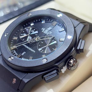 May include: A black Hublot Geneve chronograph watch with a round case, a black dial featuring silver-toned hands and markers, two sub-dials, a date window, and a black rubber strap. The bezel is secured with six visible silver-toned screws.