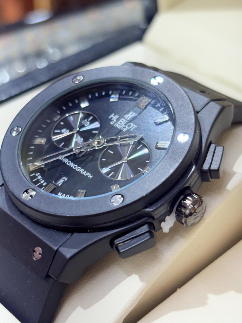 May include: A black Hublot Geneve chronograph watch with a round case, a black dial featuring silver-toned hands and markers, two sub-dials, a date window, and a black rubber strap. The bezel is secured with six visible silver-toned screws.