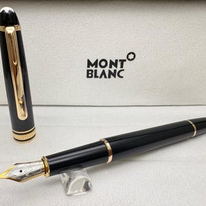 May include: A black fountain pen with gold detailing and a gold nib, displayed on a clear stand. The pen's cap is off, showing the nib. The pen is in a white box with the "MONTBLANC" logo.