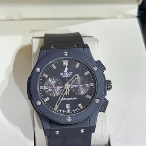 May include: A black chronograph watch with a matte finish, featuring a round dial with the text HUELOT GENEVE and CHRONOGRAPH. The watch has a black rubber strap, silver-toned hands, index markers, a date window at the six o'clock position, and six screws on the bezel.