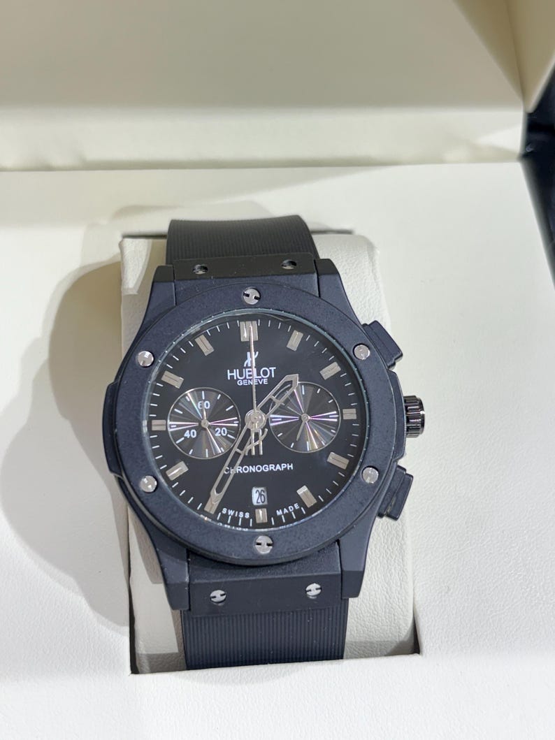 May include: A black chronograph watch with a matte finish, featuring a round dial with the text HUELOT GENEVE and CHRONOGRAPH. The watch has a black rubber strap, silver-toned hands, index markers, a date window at the six o'clock position, and six screws on the bezel.