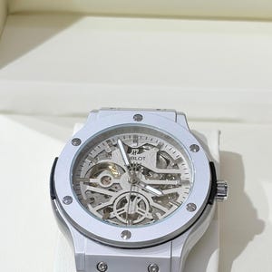 Op de afbeelding: Een zilverkleurig polshorloge met een transparante wijzerplaat, dat de interne mechanismen laat zien. Het horloge heeft een ronde bezel met zichtbare schroeven en een zwarte leren band. De merknaam "HUBLOT" is zichtbaar op de wijzerplaat.
