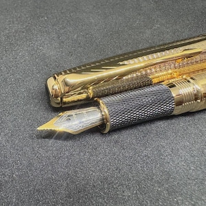 May include: A gold fountain pen with an intricate design, the cap is off and placed beside the pen. The nib is visible, with a silver and gold finish. The pen has a black textured grip. The pen is on a dark grey surface.