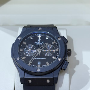 May include: A black Hublot Geneve chronograph watch with a matte black case, a black dial featuring silver-toned hour markers, three sub-dials, a date window at the six o'clock position, and a black rubber strap with a secure deployment clasp.