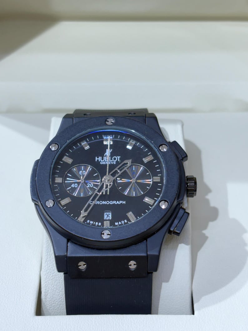 May include: A black Hublot Geneve chronograph watch with a matte black case, a black dial featuring silver-toned hour markers, three sub-dials, a date window at the six o'clock position, and a black rubber strap with a secure deployment clasp.