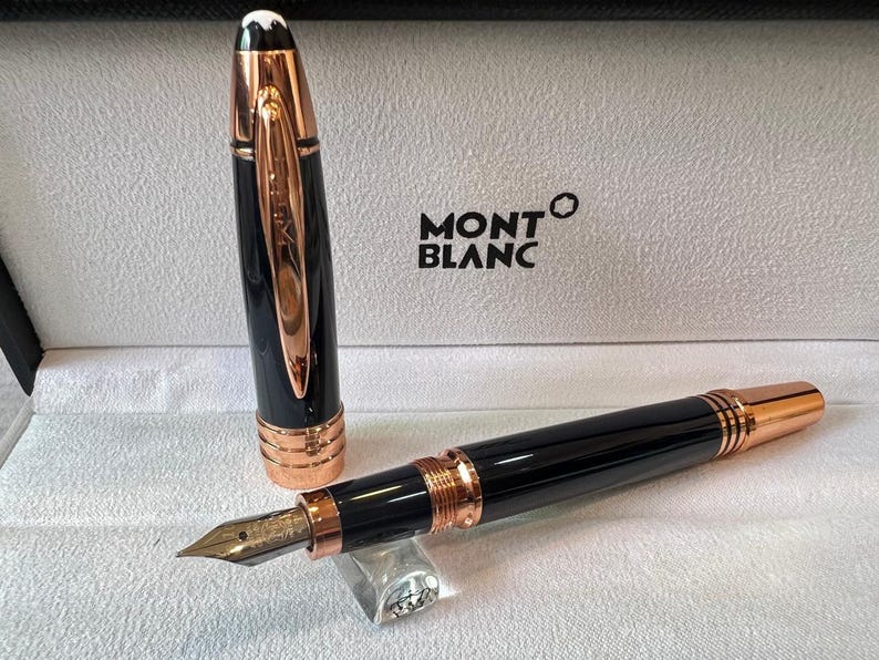 May include: A black and rose gold Montblanc fountain pen, with the cap removed. The pen is presented in its box, with the Montblanc logo. The pen features rose gold detailing, including the clip and other accents.