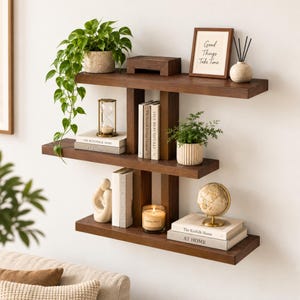 May include: A three-tiered dark brown wooden wall shelf featuring a staggered design. The shelves hold a potted trailing plant, a framed sign reading Good Things Take Time, an hourglass, books, a small green plant, a candle, and a globe.