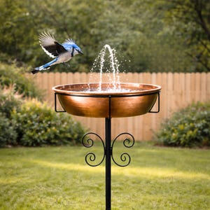 May include: A copper-colored bird bath fountain with a black metal stand. A blue jay is flying towards the water fountain. The bird bath has a decorative, scrolled base and is set in a grassy yard with a wooden fence in the background.