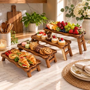 May include: A set of three tiered wooden serving risers displaying an assortment of food. The top riser holds sliced fruit and berries. The middle riser features cupcakes and cookies. The bottom riser displays savory tarts and croissant sandwiches.