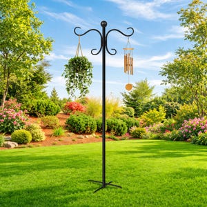 May include: A black metal garden stand with a decorative top, holding a hanging plant and wind chimes. The stand is set in a lush green lawn with a variety of colorful flowers and trees in the background.