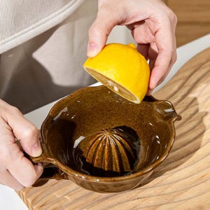 以下が含まれることがあります： 半分に切った黄色いレモンを絞るために使用されている、リブ付きの中心部を備えた茶色のセラミック柑橘類ジューサー。ジューサーにはハンドルと注ぎ口が付いています。ジューサーは木製のまな板の上にあります。