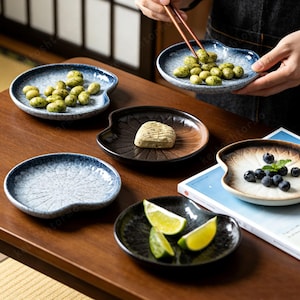May include: Several small, uniquely shaped ceramic plates in shades of blue, brown, and black. Some plates contain snacks such as green peas, blueberries, and lime wedges. The plates are arranged on a wooden table, suggesting a serving or display setting.