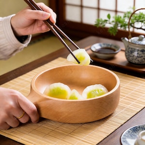 以下が含まれることがあります： 持ち手付きの木製ボウルには、いくつかの餃子が入っています。箸がボウルから餃子を一つ持ち上げています。小さなソース皿とティーポットが背景にあります。ボウルは竹のマットの上に置かれています。