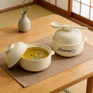 以下が含まれることがあります： 蓋とスプーンが付いた、オフホワイトのセラミック製スープボウルが2つ。1つのボウルは開いており、スープと麺が入っています。もう1つのボウルには「Hanna」という名前とハートのマークが付いています。ボウルは木製のテーブルの上の茶色のプレースマットの上にあります。
