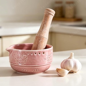 May include: A pink ceramic mortar and pestle set with a wooden pestle. The mortar has embossed cat designs and a spout. A whole garlic bulb and a clove are next to the mortar.