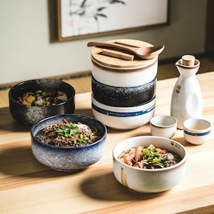 May include: A selection of Japanese ceramic bowls and serving dishes. The set includes bowls with food, a stack of bowls with a wooden lid, a sake bottle, and small cups. The bowls have blue, black, and white designs.