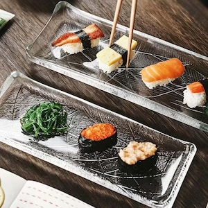 May include: Two rectangular clear glass serving plates with a textured leaf-vein pattern. The top plate holds four pieces of nigiri sushi, including eel, tamago, and salmon, with wooden chopsticks. The bottom plate holds seaweed salad and two gunkan maki.