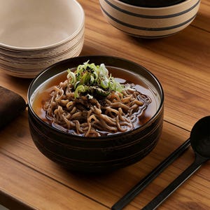 以下が含まれることがあります： そばの麺と出汁が入った黒い器のクローズアップ。ネギとゴマがトッピングされています。器はテクスチャ加工が施されています。箸とスプーンが木製の表面に置かれています。
