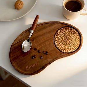 以下が含まれることがあります： 木製のサービングトレイ、コースターとスプーン付き。トレイは茶色で、「Emma」という名前が刻まれています。木製の柄が付いたステンレス製のスプーンがトレイの上に置かれ、コーヒー豆も添えられています。コーヒーカップとクルミの入ったお皿もあります。